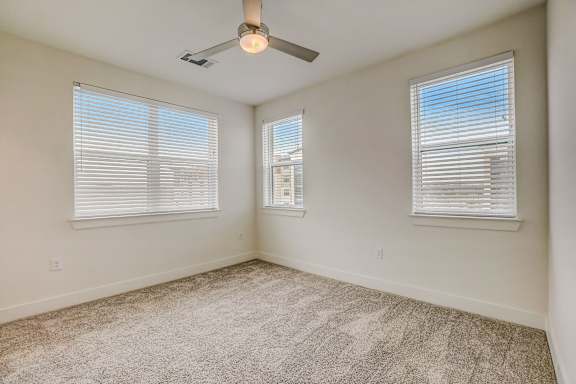 A room with a carpeted floor, a ceiling fan, and two windows with blinds.