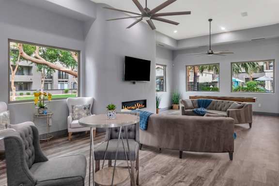 A living room with a grey couch, a white armchair, a grey chair, a round table, a television, a fireplace, and a ceiling fan.