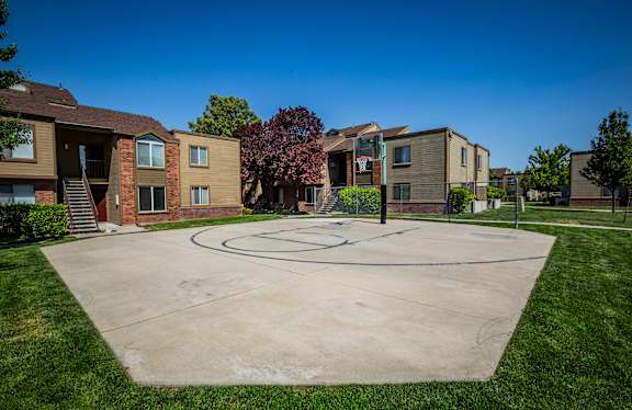 A basketball court is located in the middle of a grassy area in front of apartment buildings.