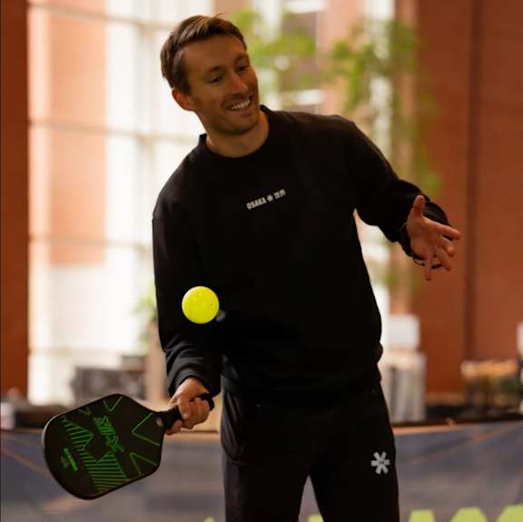 A man in a black sweatshirt and shorts is playing with a yellow ball and a paddle.