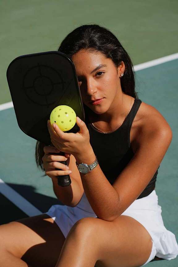 A woman is sitting on a tennis court holding a tennis ball and a racket.
