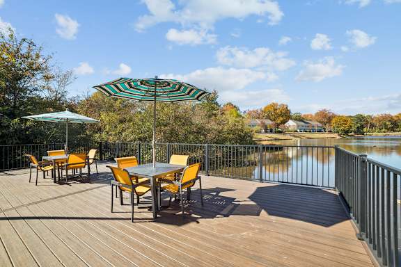 A table with chairs and an umbrella on a wooden deck overlooking a lake at Lakecrest Apartments, PRG Real Estate Management, Greenville, SC 29615