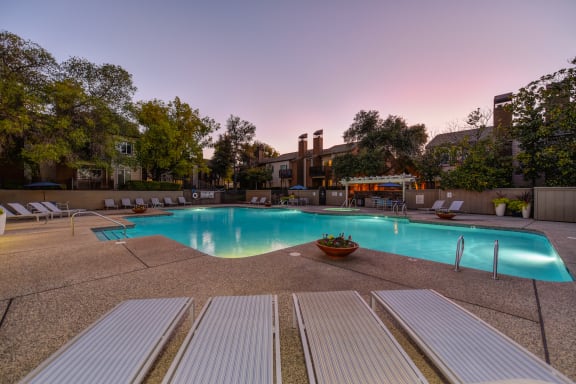 Swimming pool at dusk. The pool lights are on and pool is illuminated.  There are lounge chairs and umbrellas all around the pool area.at Folsom Ranch, Folsom