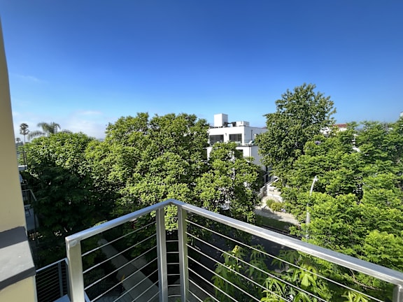 A balcony with a metal railing overlooks a lush green area with trees and a white building in the distance.