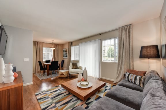 Dining Area and Living Room with Wood Inspired Floors,  Gray Sofa, Short Wood Table, Open Window Blinds, Lamp, Dining Room Table and Chairs at Addison Ranch Apartments, California