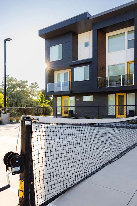 A tennis court with a black net in front of a modern building with balconies.