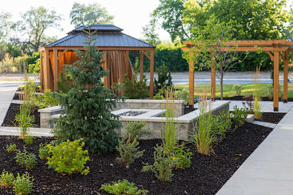 A gazebo is surrounded by a garden with a wooden pergola overhead.