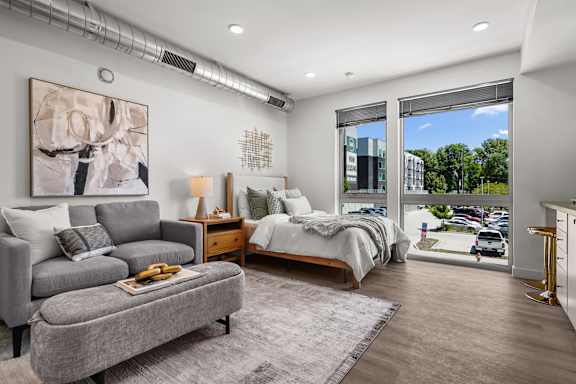 A bedroom with a bed, a couch, a nightstand, and a window overlooking a parking lot.