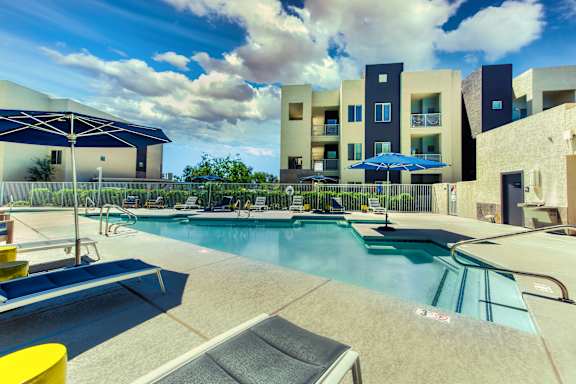 A swimming pool with a yellow sun lounger and a blue umbrella. at Craft at Gilbert and Baseline Apartments, Mesa, Arizona