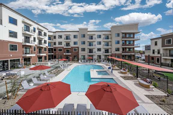 A large swimming pool surrounded by red umbrellas and chairs in front of apartment buildings.
