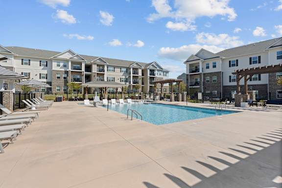 A large swimming pool surrounded by sun loungers in front of apartment buildings.