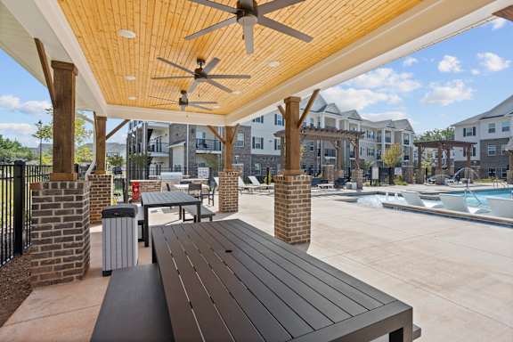 A wooden bench is in the foreground of a patio with a ceiling fan and a pool in the background.