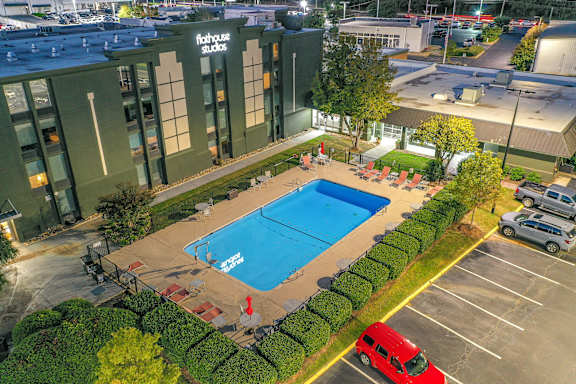 A swimming pool is surrounded by a hedge and a red van is parked in the parking lot.