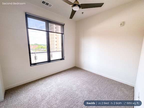 A second bedroom with a carpeted floor, a ceiling fan, and a large window overlooking a cityscape.