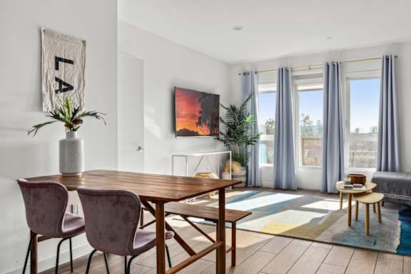 Dining area with a wooden table and chairs and living room view with large windows and curtains.