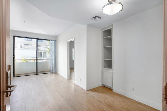 A room with a sliding glass door leading to a balcony, a wooden floor, and a white wall with a built-in shelf.