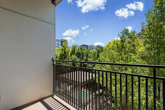 A balcony with a black railing overlooks a pool and green trees.