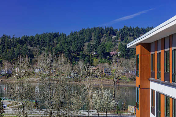 A building with a brown and white exterior sits in front of a body of water and a wooded area.