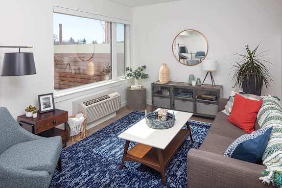 A living room with a grey couch, a grey armchair, a wooden coffee table, a round mirror, a lamp, a vase, and a potted plant.