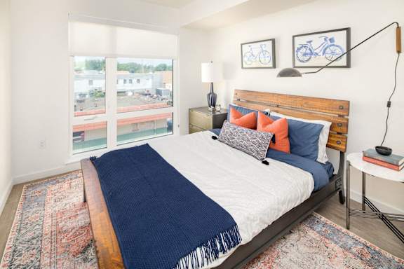 A bedroom with a bed, a nightstand, and a window overlooking a parking lot.