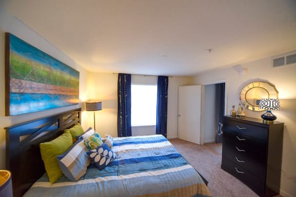 Spacious bedroom with carpet flooring, walk-in closet, staged with accent wall, dresser, side table and desk lamp