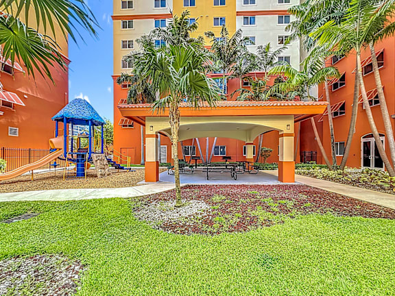 A playground area with a slide and a gazebo is surrounded by palm trees and a building.