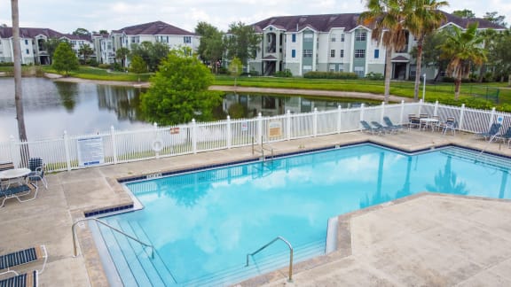 Community pool with sundeck, lounge chairs, with lake view surrounded by white metal fence with palm trees and building exteriors and lake in the background
