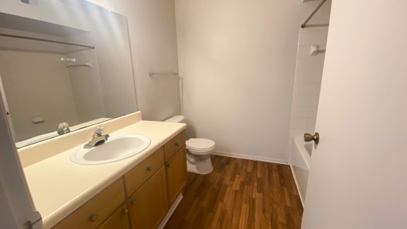 Bathroom with wood floors, large mirror toilet, shower tub combo with tile back splash, towel bar and brown wood cabinets