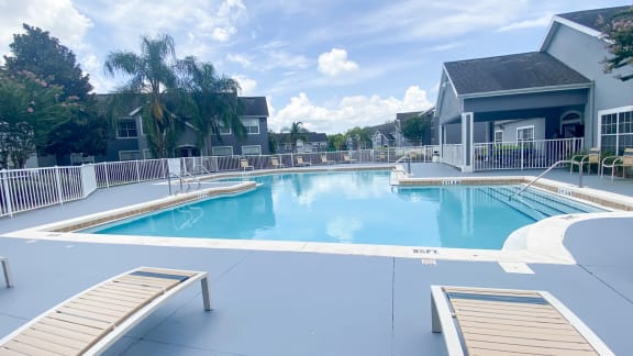 Community pool with sundeck, lounge chairs, with lake view surrounded by white metal fence with palm trees and building exteriors, palm trees, and trees in the background