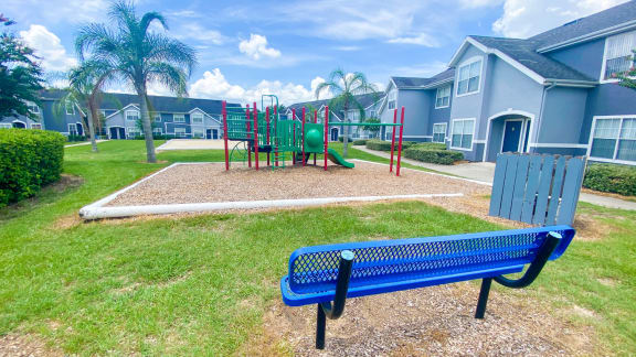 Green and Red Playground set in a bed of mulch with buildings and palm tree in the background, and a  blue bench in the front