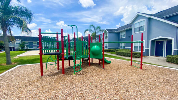 Green and red Playground set in a bed of mulch with buildings and palm tree in the background