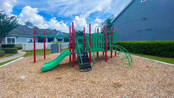 Green and Red Playground set in a bed of mulch with buildings and palm tree in the background