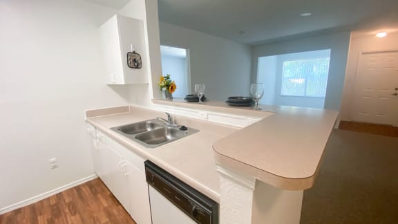 Kitchen featuring white cabinets, white dishwasher, brown vinyl counter top, with place setting plate, bowl and long stem wine glass along with flowers on breakfast bar top, with view of living room