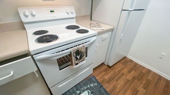 Kitchen featuring white cabinets, white appliances, tan countertop, and electric stove top with coils, wood style flooring, with floor mat and hand towels as décor