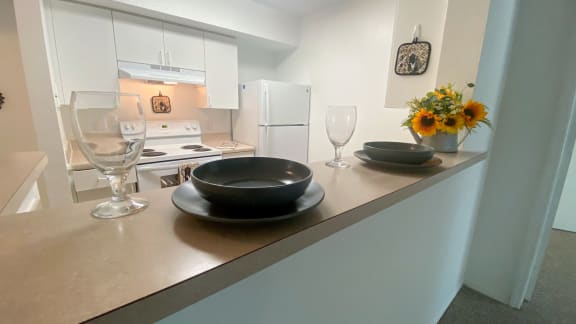 Kitchen featuring white cabinets, white appliances, brown vinyl  counter top, vent hood over electric stove, with place setting plate, bowl and long stem wine glass along with flowers on bar top