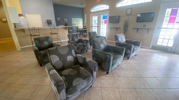 Six arm chairs in clubhouse with two flat screen televisions to the side of them, and the kitchen and dinning room in the back ground on a tile floor, two tone paint