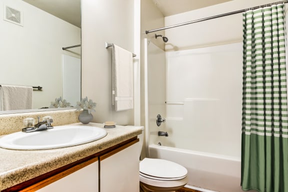A virtually staged bathroom with white walls, hardwood style flooring, a single toilet, towel bar, a single sink with storage below and a mounted mirror behind the counter/sink area. There is a tub/shower combo with a green and cream checkered shower curtain, a cream towel, and a small vase with flowers on the bathroom countertop.