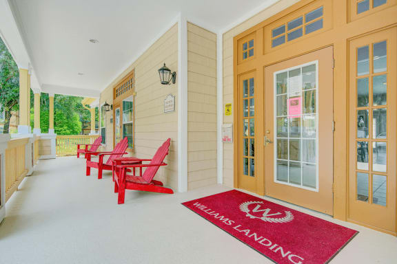 Exterior clubhouse entrance with covered porch featuring red adirondack chairs, red welcome mat, and tan door frame entrance