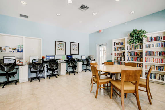 Business center with multiple computers and chairs, a wooden table with chairs and library with book shelves.