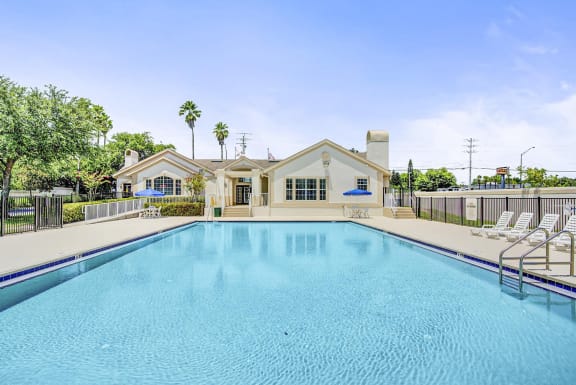 Swimming pool and sundeck with tables, chairs, and loungers. The perimeter fence is lined with trees and the exterior of the clubhouse.