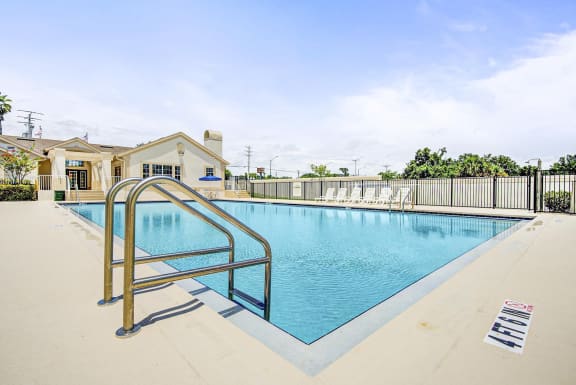 Swimming pool and sundeck with tables, chairs and loungers. The perimeter fence is lined with trees and the exterior of the clubhouse.