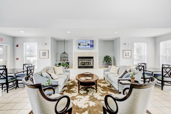 Clubhouse lounge with couches, chairs, coffee table, tiled flooring, tv mounted above fire place with two table and chair sets on either side of lounge area.