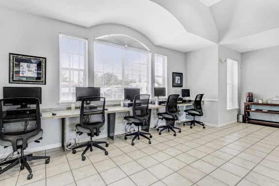 Business center with five desktop computers with flat-screen monitors, four chairs, a printer, and large windows with a coffee bar in the back