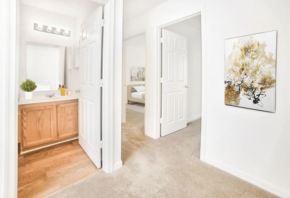 Carpeted bedroom looking towards bathroom with wood flooring and vanity. Virtually staged bathroom with décor and gold painting