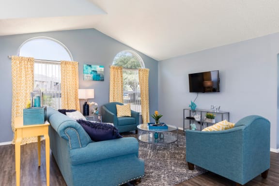 Clubhouse interior with hardwood style flooring, blue couch and two armchairs. Lounge area in clubhouse with blue and yellow decorations. Blue couch, yellow curtains, mounted tv, and glass table.