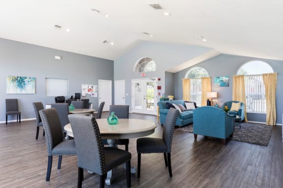 Clubhouse interior with hardwood style flooring, blue couch and two armchairs. Two sets of round tables with chairs and leasing desk in the background