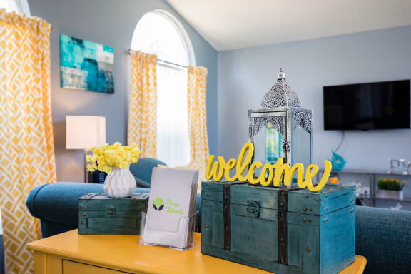 Lounge area in clubhouse with blue and yellow decorations. Blue couch, yellow curtains, mounted tv, and welcome sign.