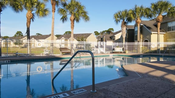 Community pool with sundeck, lounge chairs, with lake view surrounded by white metal fence with palm trees and building exteriors in the background
