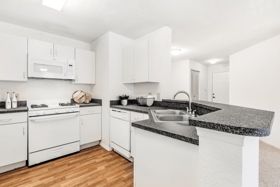 Virtually staged kitchen with white appliances, wood flooring, gas stove, white cabinetry, gray speckled countertop