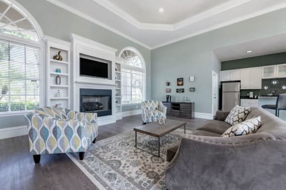 Clubhouse Seating Area with Large Windows on either side of Fireplace with TV and Shelves Surrounding Fireplace Multi Colored Accent Chairs Grey Couch and Clubhouse Kitchen with White Cabinets and Stainless Steel Appliances